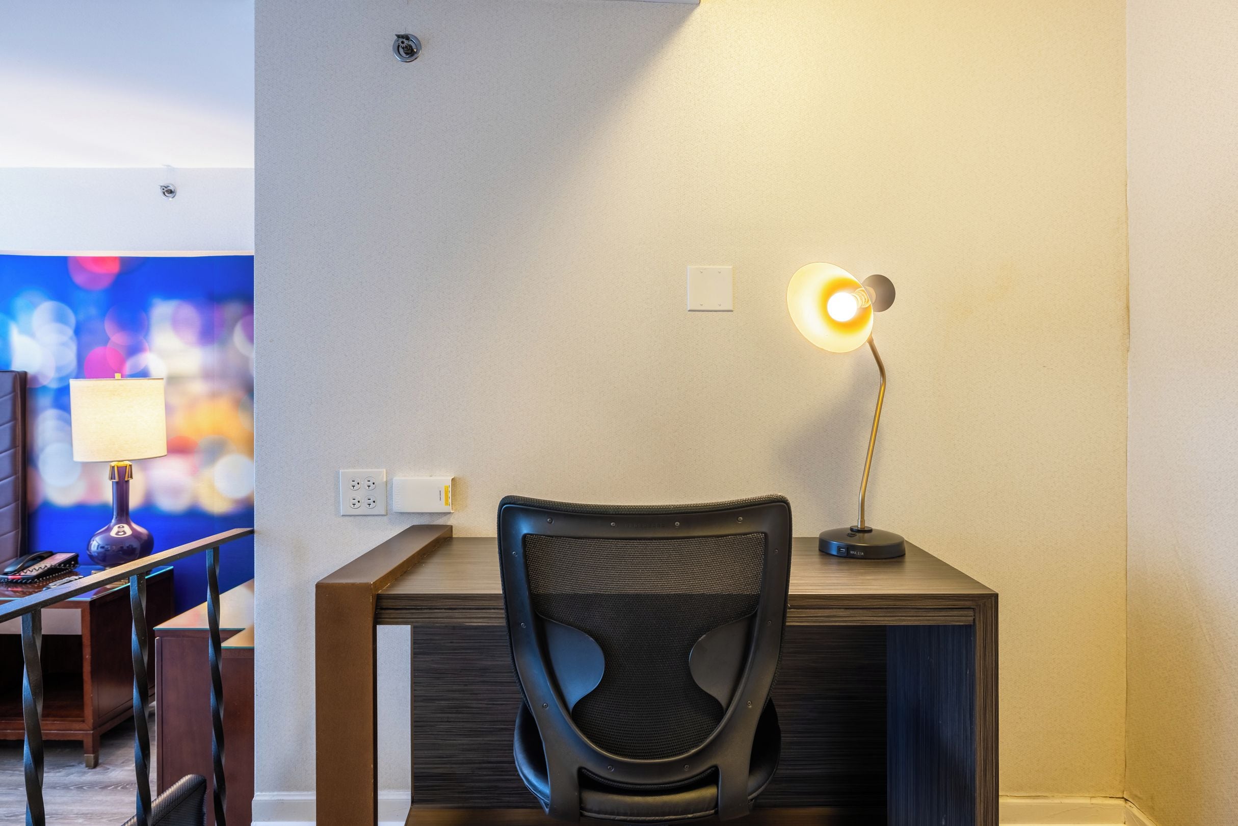 A hotel room with a desk, a chair, a lamp, and a wall socket.