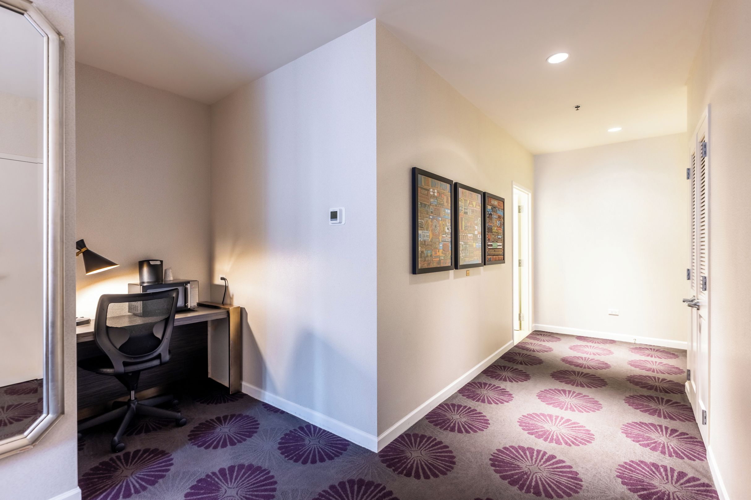 A hallway with a carpeted floor, a desk with a chair, and two framed pictures on the wall.