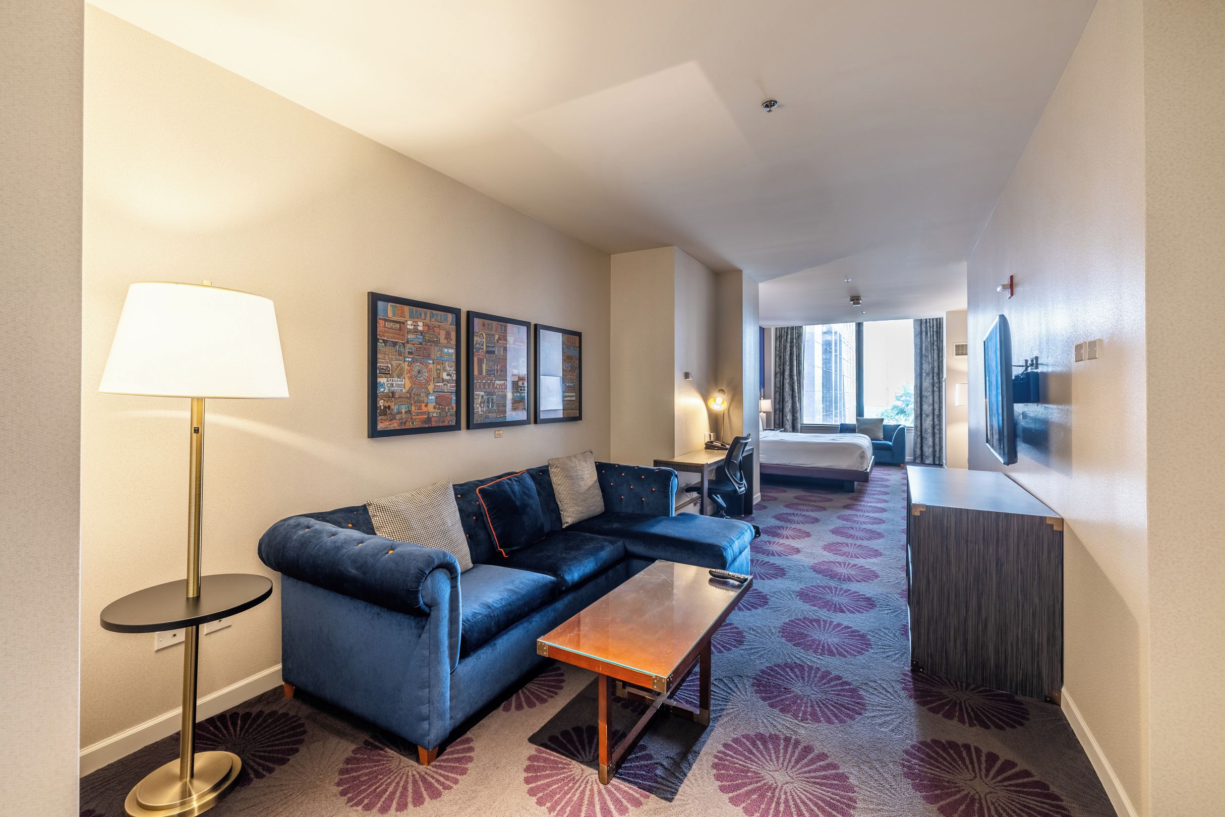 Ein Hotelzimmer mit Sofa, kleinem Tisch, Lampe, Bett, Schreibtisch und einem großen Fenster.