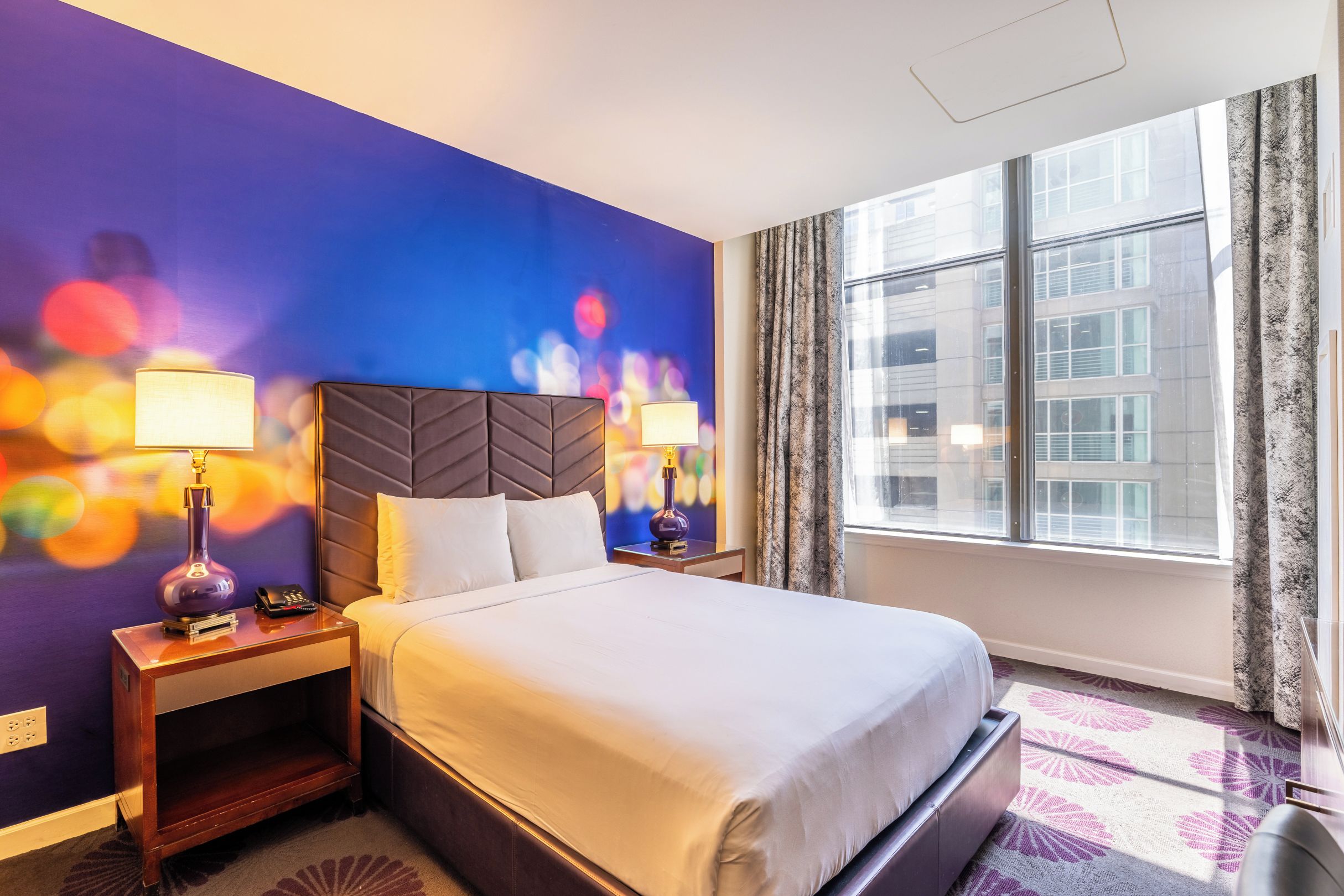 A hotel room with a large bed, two bedside tables with lamps, a wall with a colorful mural, a window with curtains, and a view of buildings outside.