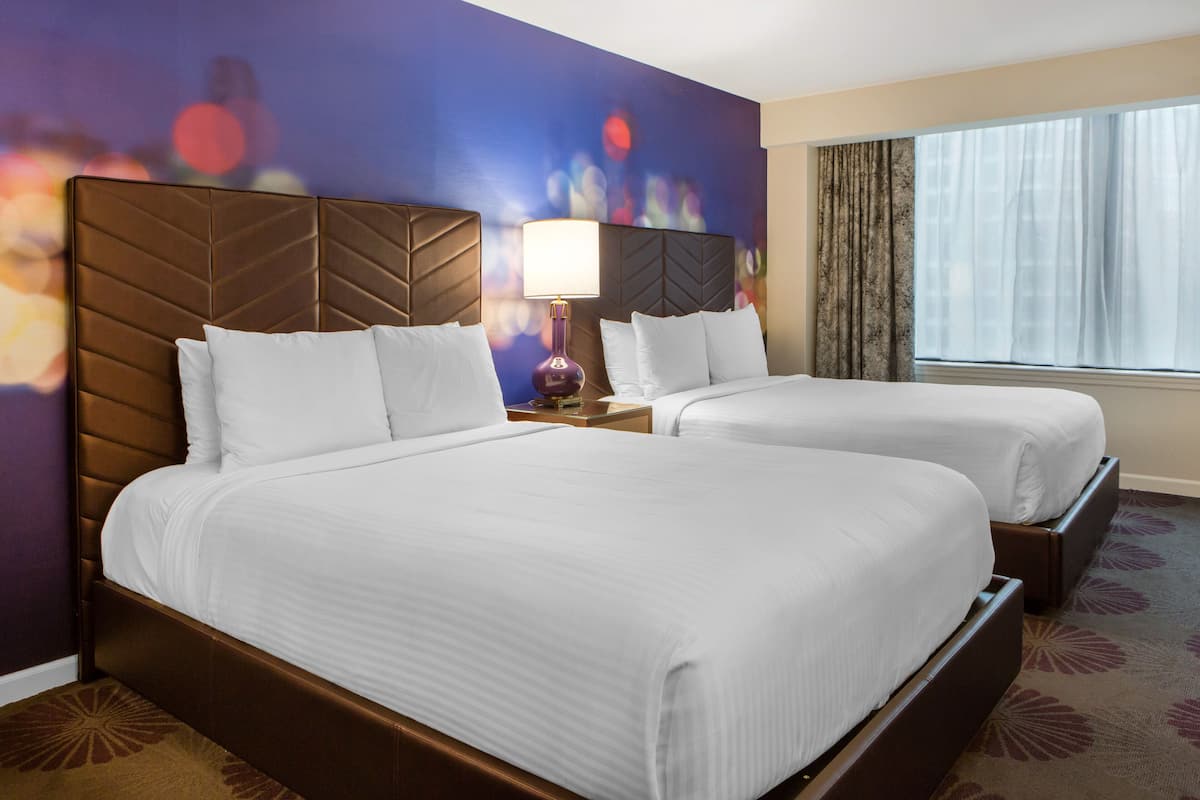 A hotel room with two beds, a nightstand with a lamp, a large window with curtains, and a wall with a bokeh light effect.