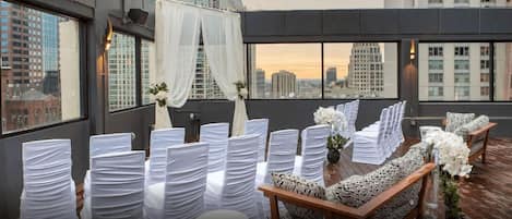 Un espacio en la azotea con sillas blancas y un sofá, cubierto con telas blancas y arreglos florales, con vistas a un paisaje urbano.