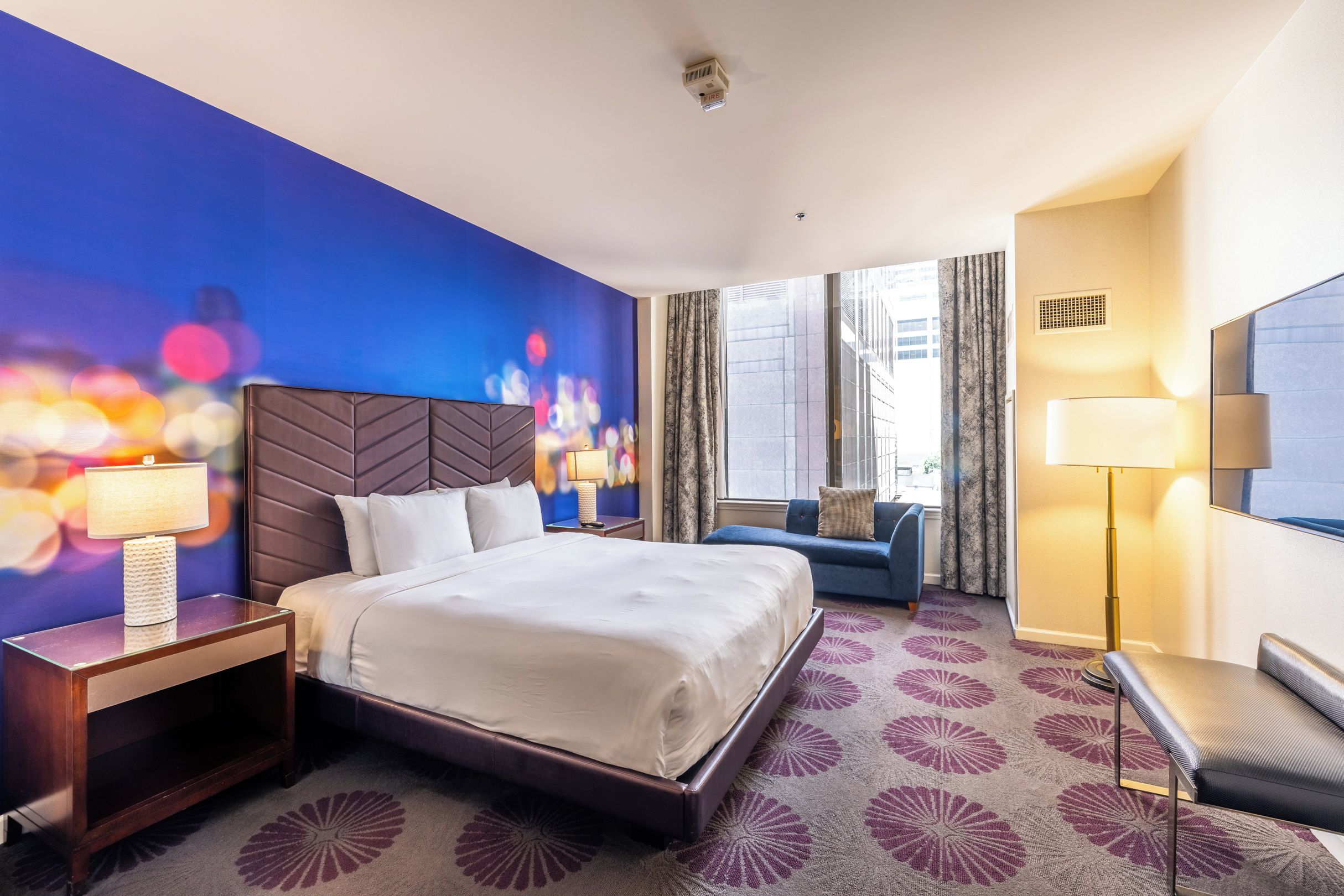 A hotel room with a large bed, a bedside table with a lamp, a seating area with a sofa, and a view of the city through the window.