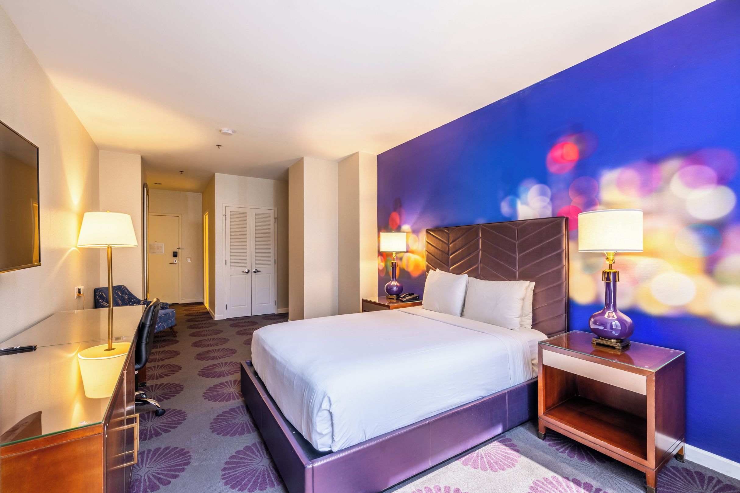 A hotel room with a bed, two bedside tables with lamps, a desk with a chair, and a mirror.