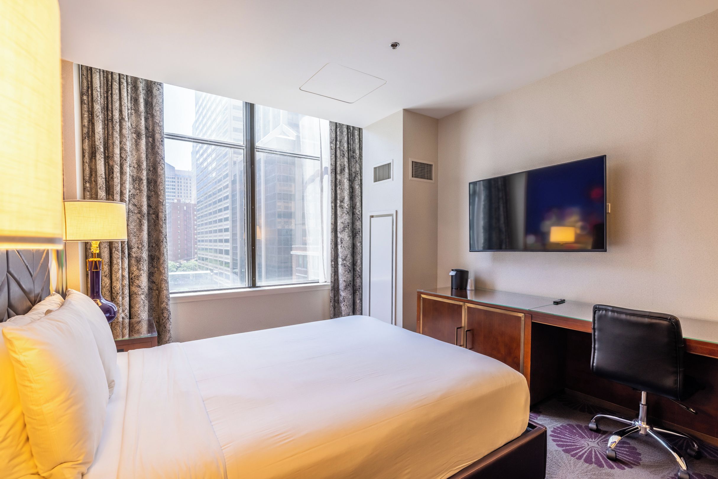 A hotel room with a large bed, a desk, a chair, a television, and a window with curtains.