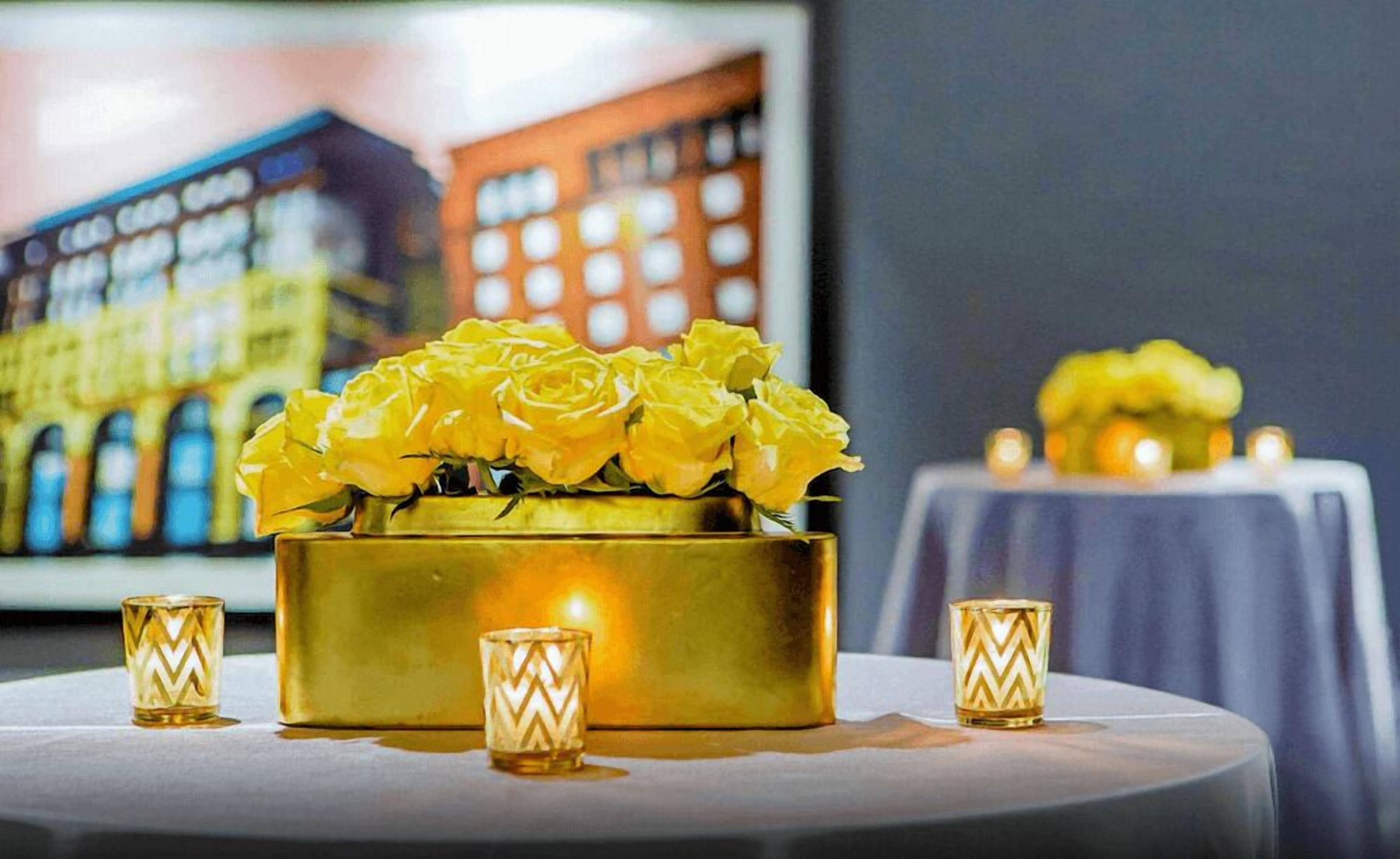 A table with a gold vase containing yellow roses, flanked by two small, patterned candle holders.