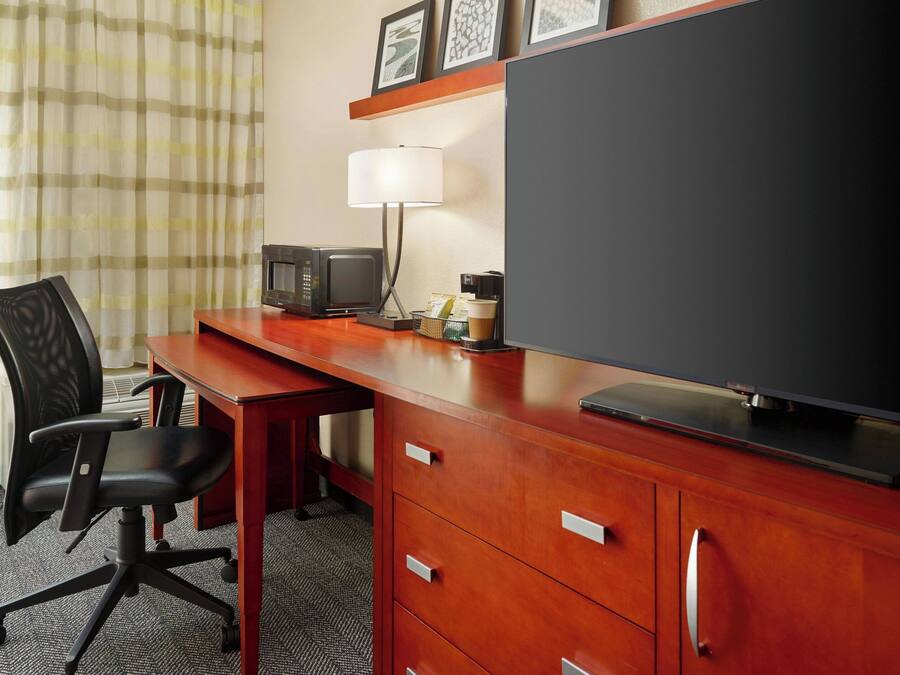 A hotel room with a wooden desk, a black office chair, a television, a microwave, a lamp, and a small coffee maker.