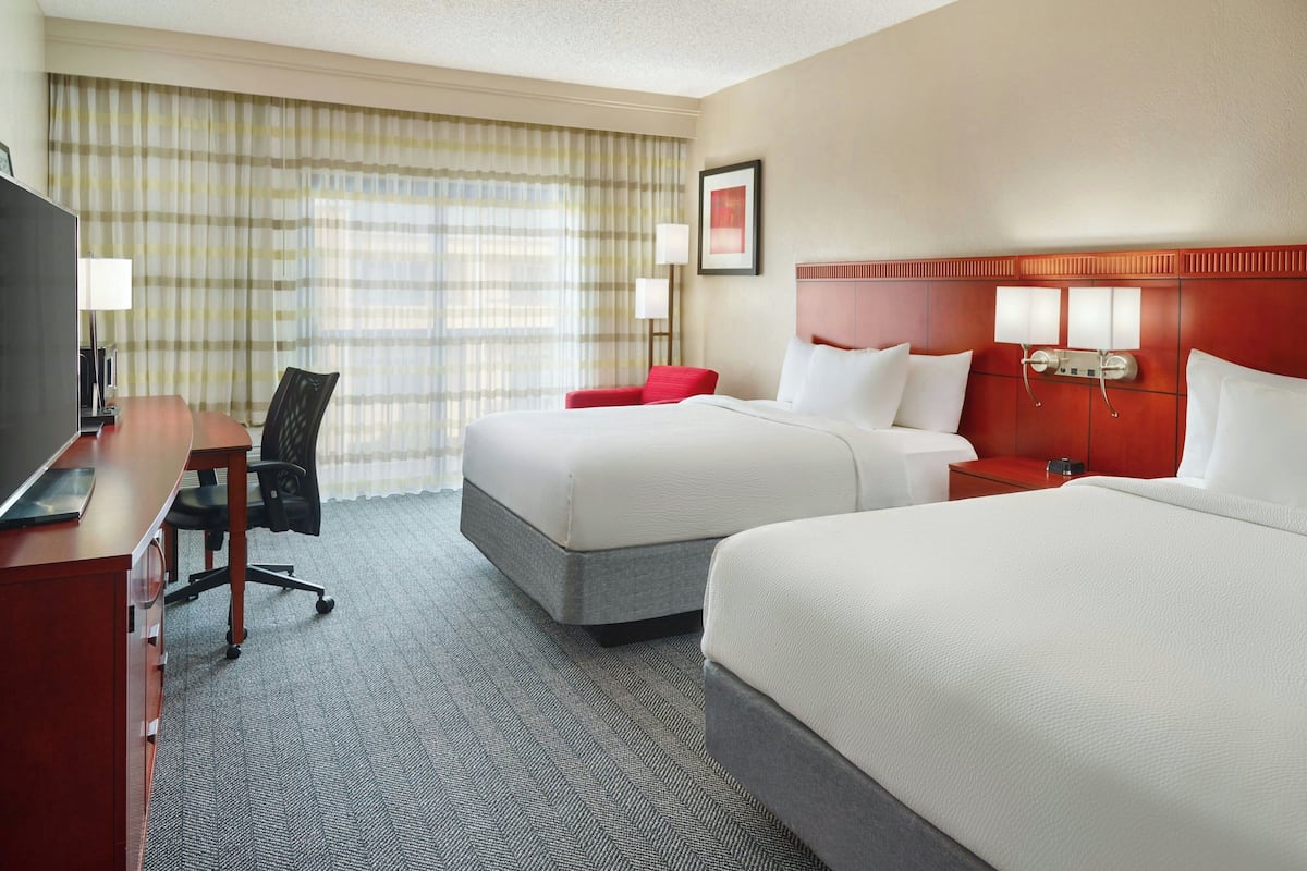 A hotel room with two beds, a desk, a chair, a TV, and a large window with curtains.