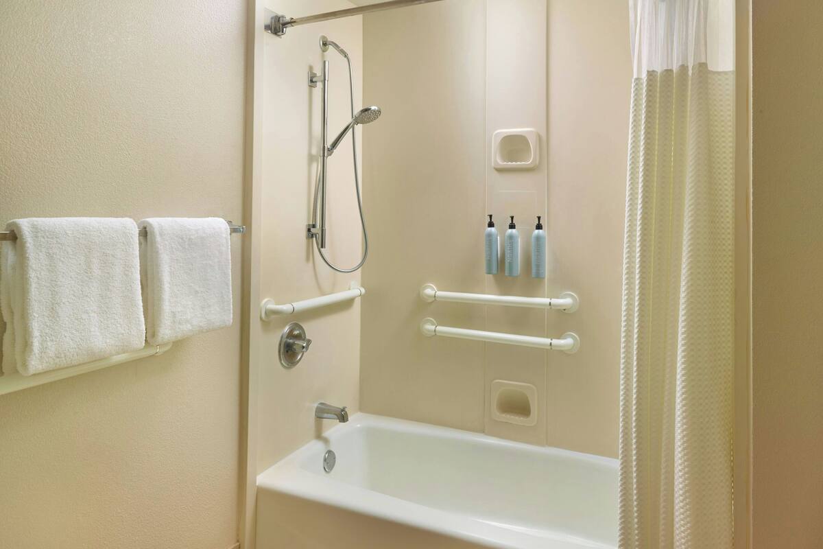 A bathroom with a bathtub, a shower, and handrails. There are towels hanging on a rack and bottles of shampoo and conditioner on the wall.