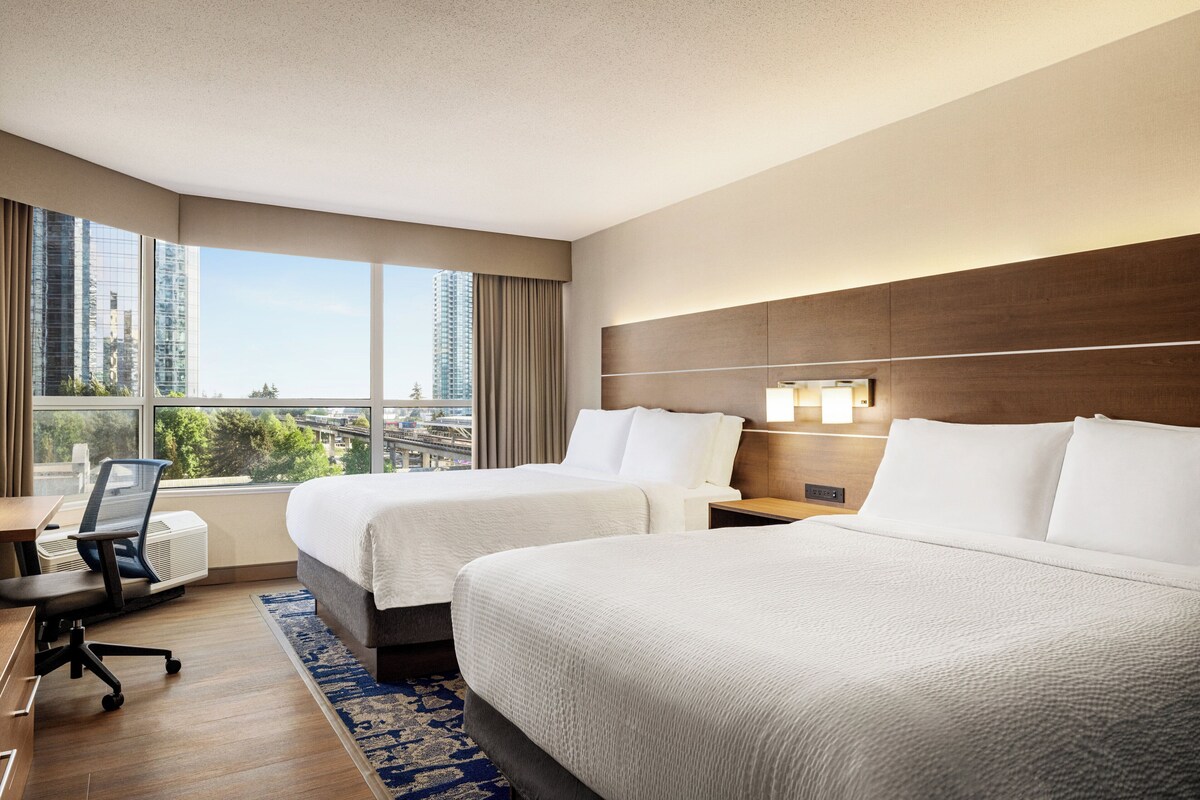 A hotel room with two beds, a desk, an air conditioning unit, and a large window with a city view.