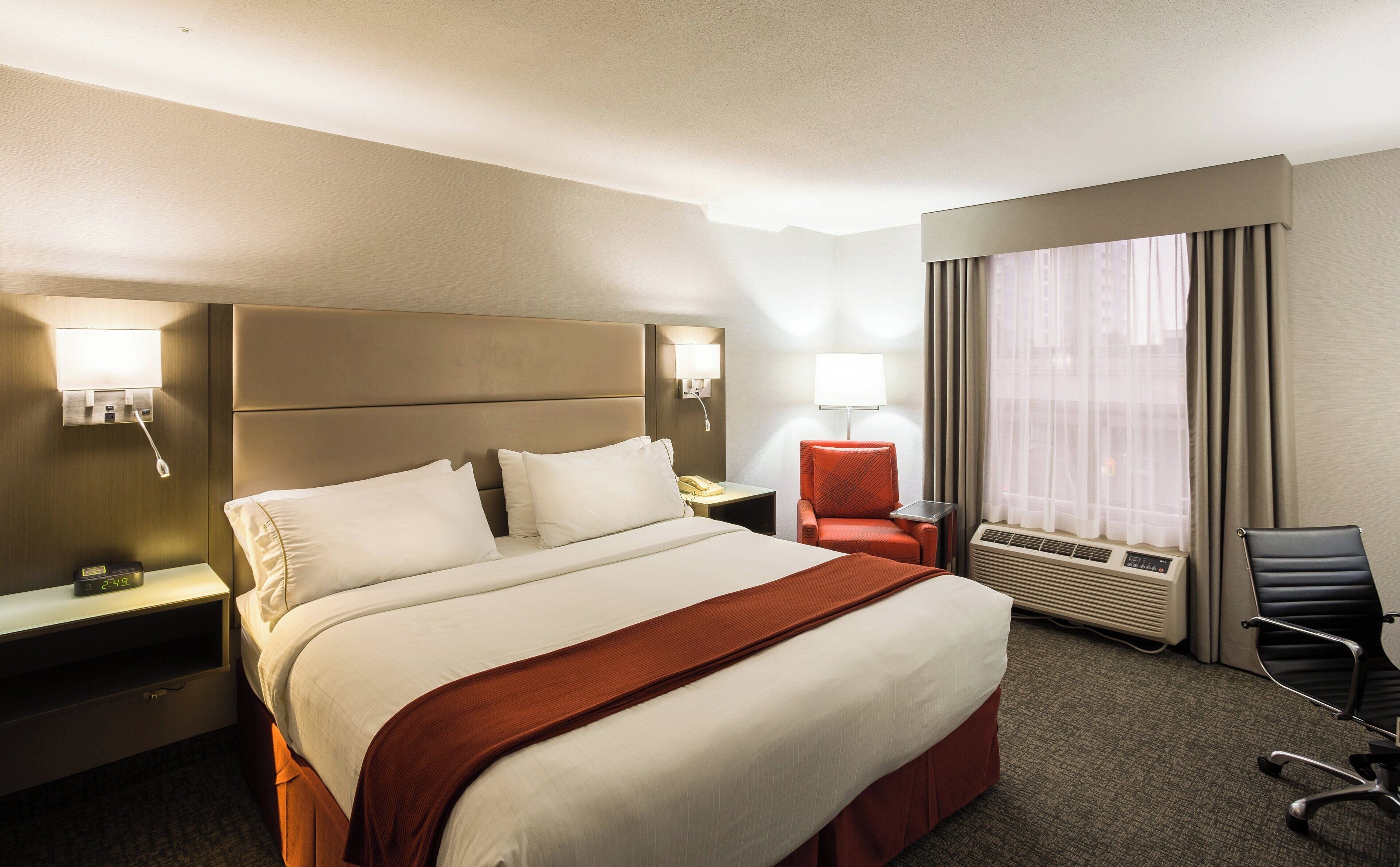 A hotel room with a large bed, bedside lamps, a desk with a telephone, a chair, and an air conditioning unit.