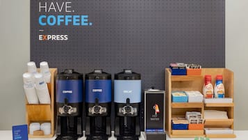 A coffee station with three coffee dispensers, a hot water dispenser, and a coffee maker. There are also coffee pods, milk cartons, and a sign that says "Start the daily grind."