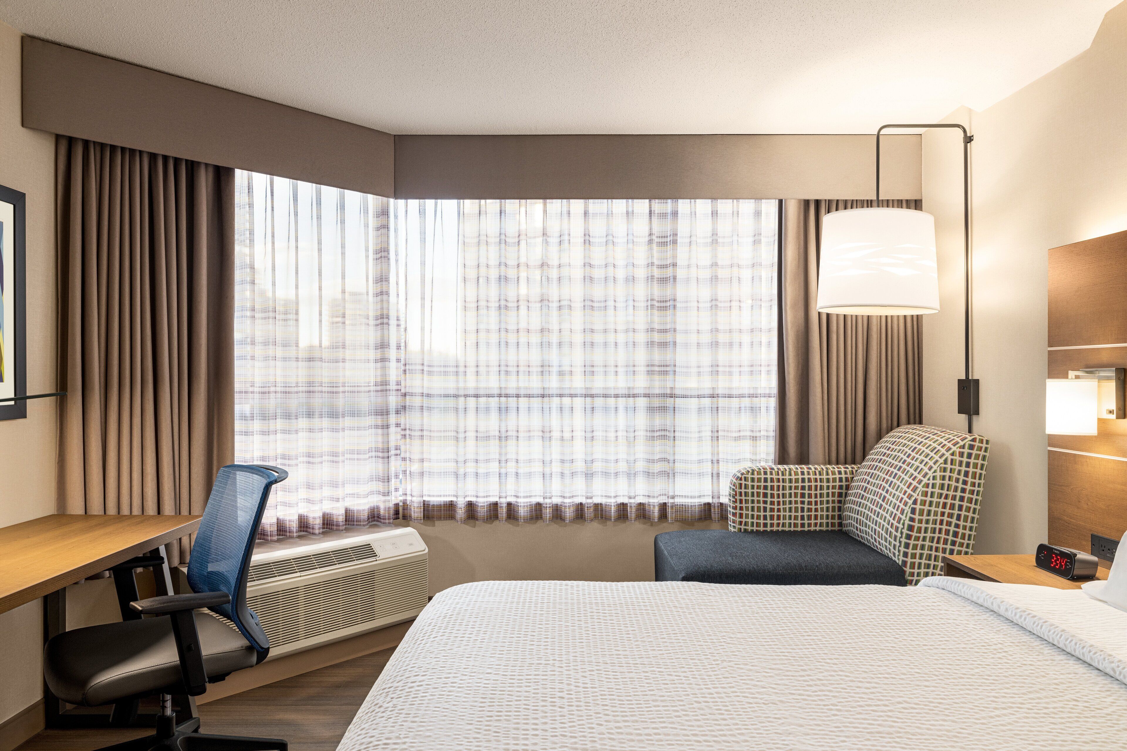 A hotel room with a bed, a desk, a chair, a lamp, and a window with curtains.