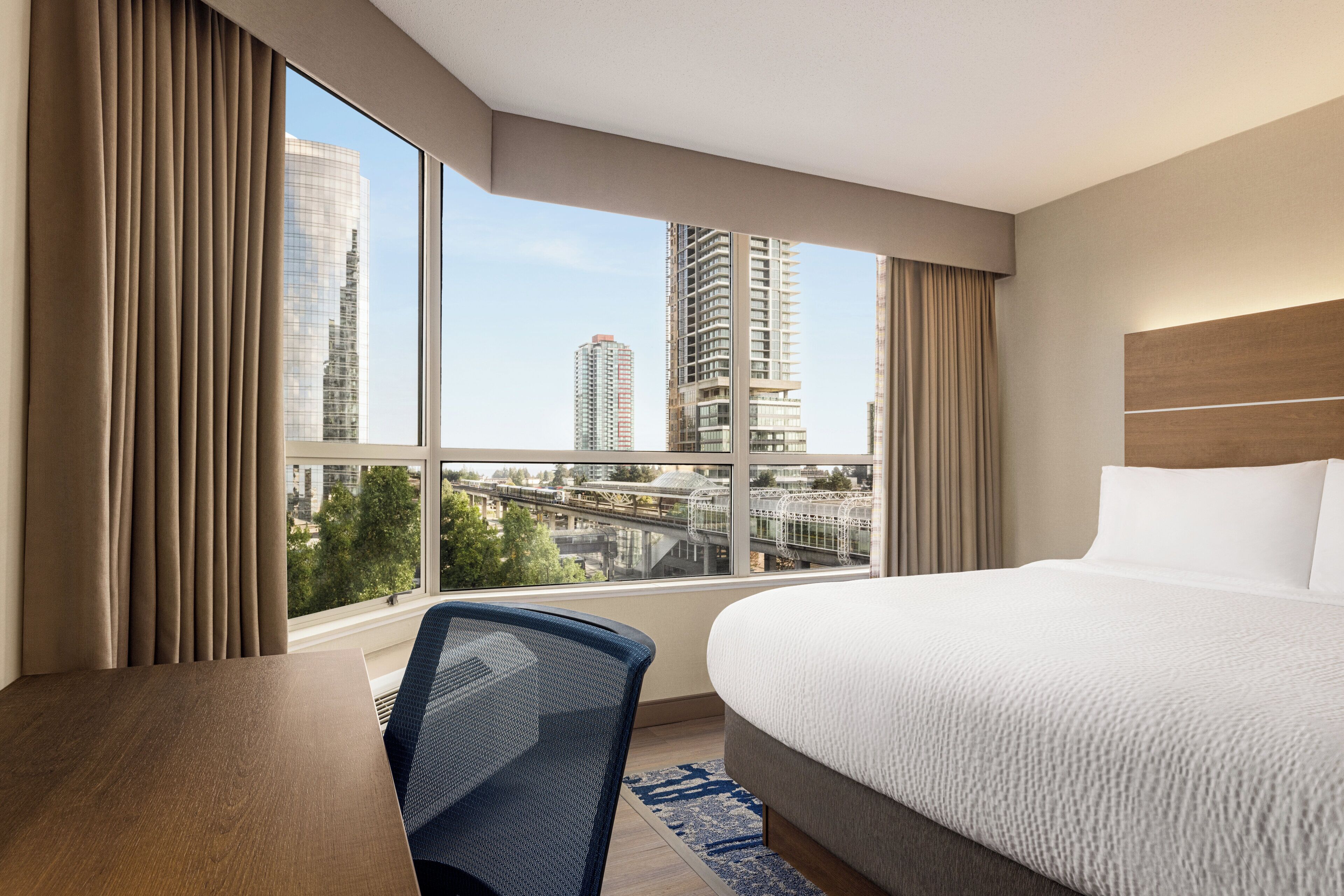 Une chambre d’hôtel avec un grand lit, un bureau et une chaise. La fenêtre offre une vue sur de hauts immeubles et un pont.