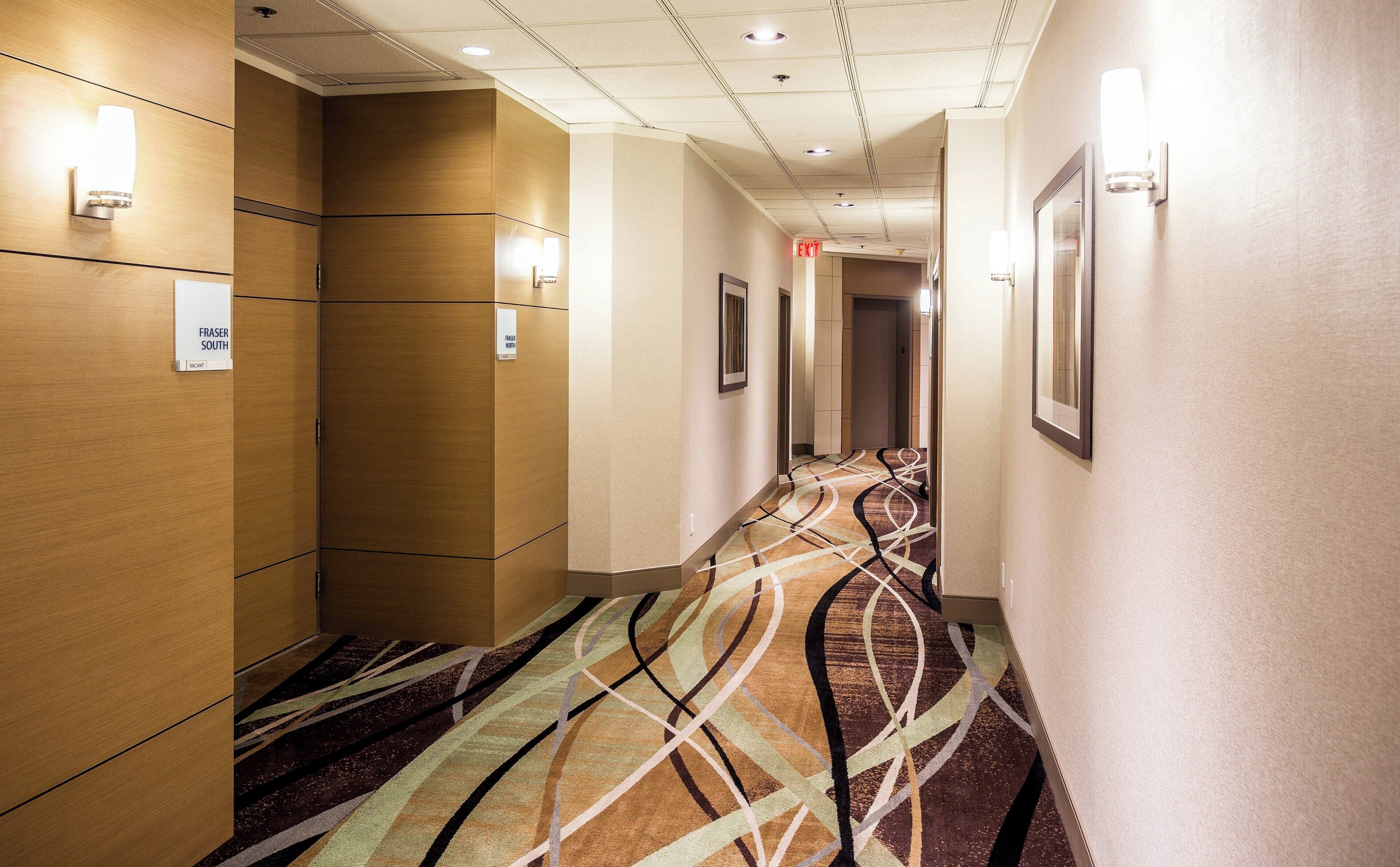 Un pasillo de hotel con paneles de madera, alfombra estampada e iluminación empotrada.