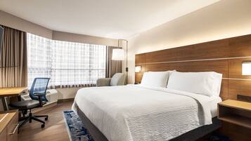 Habitación de hotel con una cama grande, un escritorio con silla, un ventanal con cortinas y una mesita de noche con lámpara.