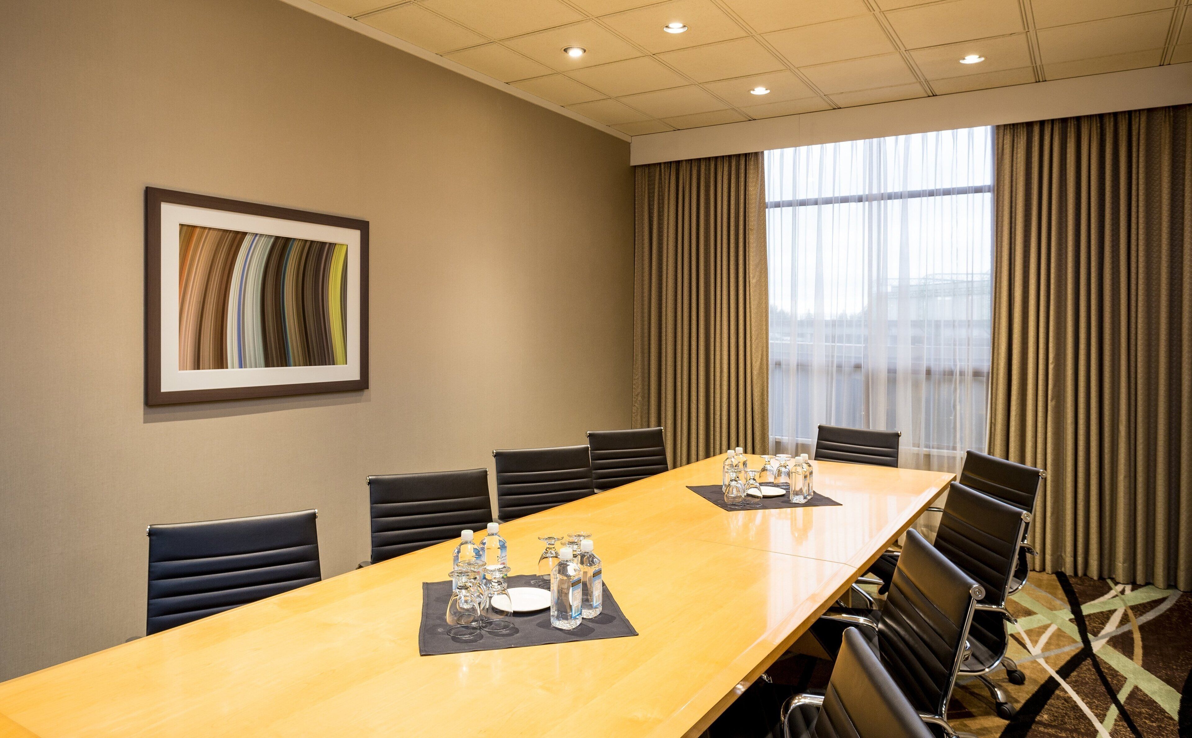 A conference room with a long table, chairs, a framed picture on the wall, and a window with curtains.