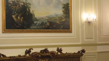 Una habitación con un cuadro clásico de grandes dimensiones, paneles de pared ornamentados, una araña y filas de sillas rojas.