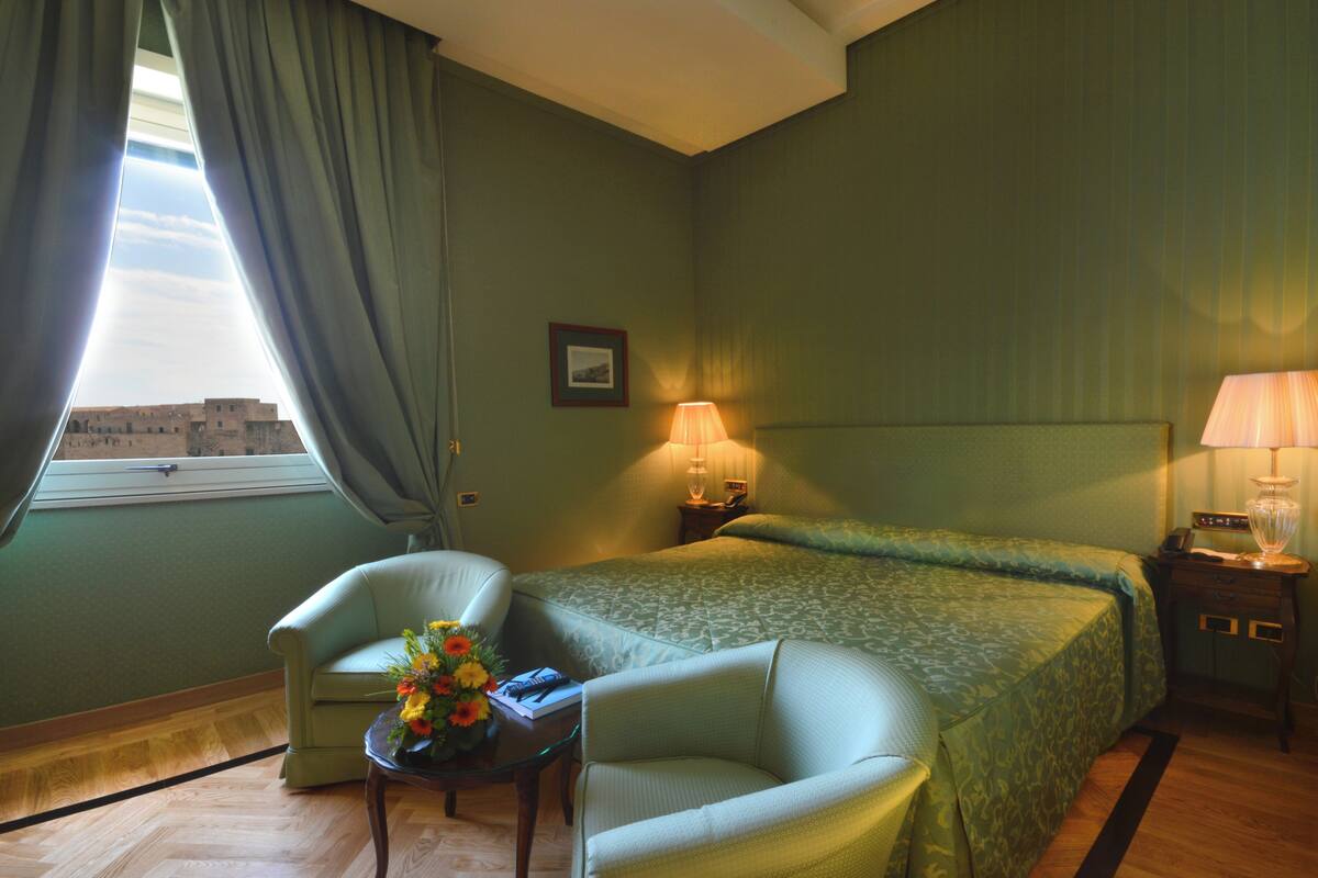 Ein Hotelzimmer mit einem Bett, zwei Sesseln, einem kleinen Tisch mit Blumenvase, einer Lampe und einem fenster mit Vorhängen.