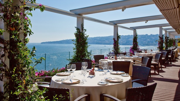 Un espace de restauration en plein air, avec des tables dressées pour un repas, offrant une vue sur une étendue d’eau et une côte lointaine.