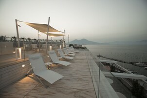 A rooftop terrace with white lounge chairs and a canopy, overlooking a marina and distant mountains.