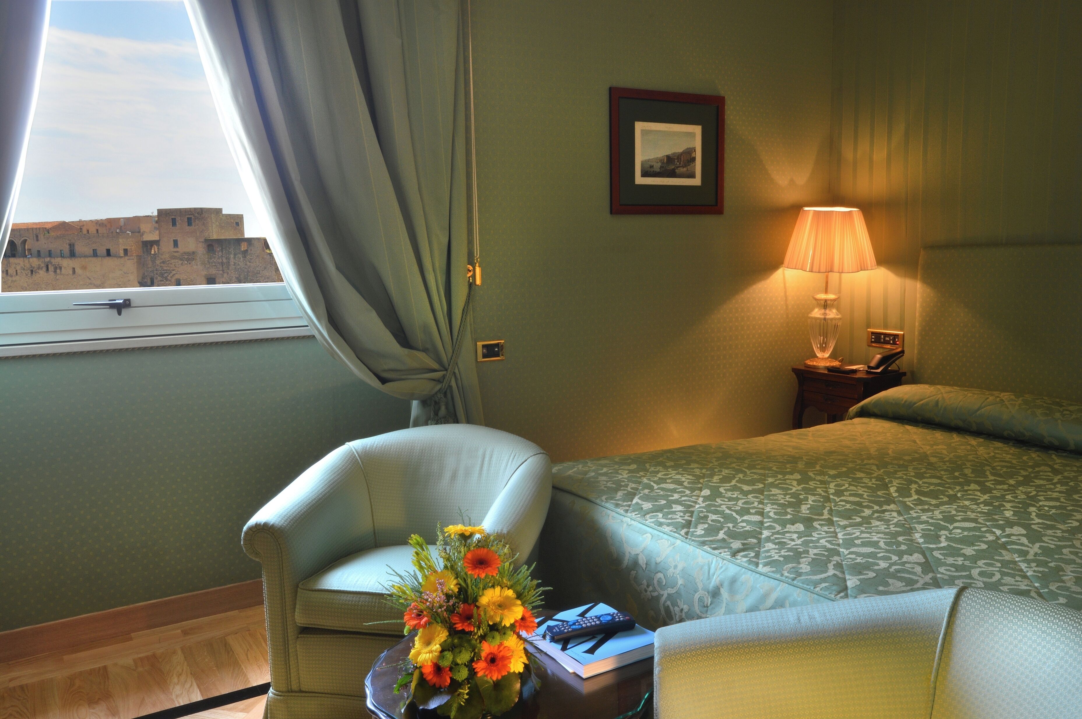 Una habitación de hotel con una cama, una silla, una mesa con un florero y una lámpara, además de un cuadro en la pared.