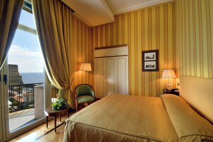 Une chambre d’hôtel avec un grand lit, deux lampes de chevet, une chaise, une petite table agrémentée d’un arrangement floral, une armoire et un balcon avec vue sur la mer.