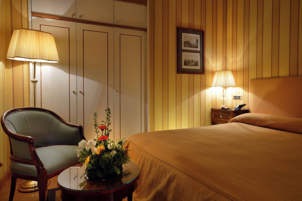 Ein Hotelzimmer mit Bett, Sessel, Tisch mit Blumenvase, Nachttisch mit Lampe und einem gerahmten Bild an der Wand.