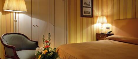 A hotel room with a bed, a chair, a table with a vase of flowers, a nightstand with a lamp, and a framed picture on the wall.