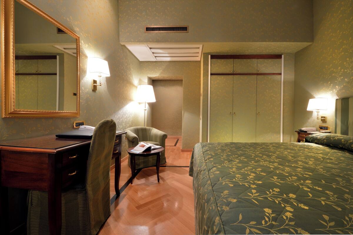 Ein Hotelzimmer mit einem Bett, einem Schreibtisch mit Stuhl, einem Spiegel und zwei Nachttischlampen.