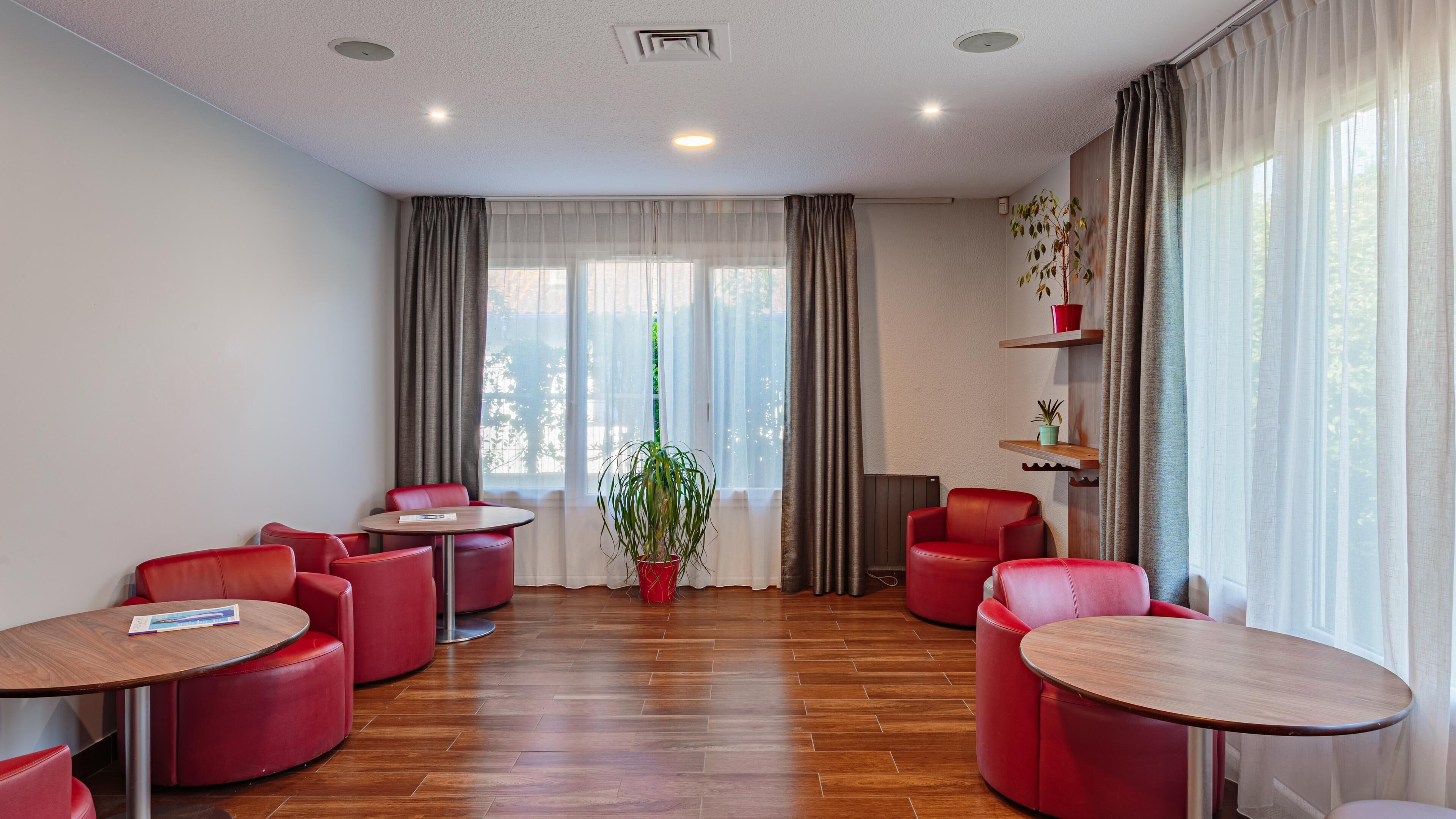A hotel room with a wooden floor, red leather chairs, a central table, and a potted plant.