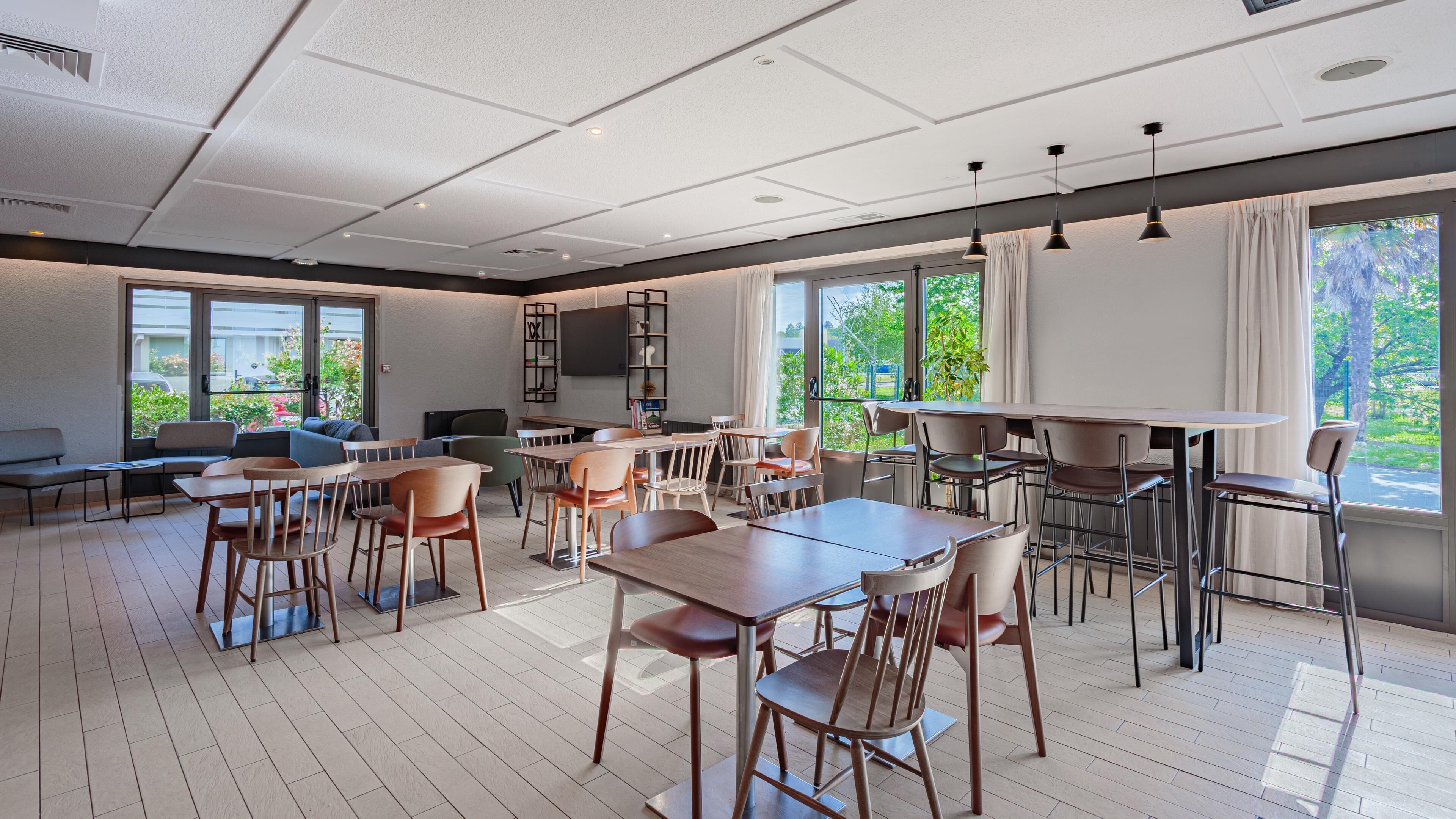 Un espace intérieur moderne avec des tables et des chaises en bois, de grandes fenêtres avec des rideaux et un téléviseur à écran plat fixé au mur.