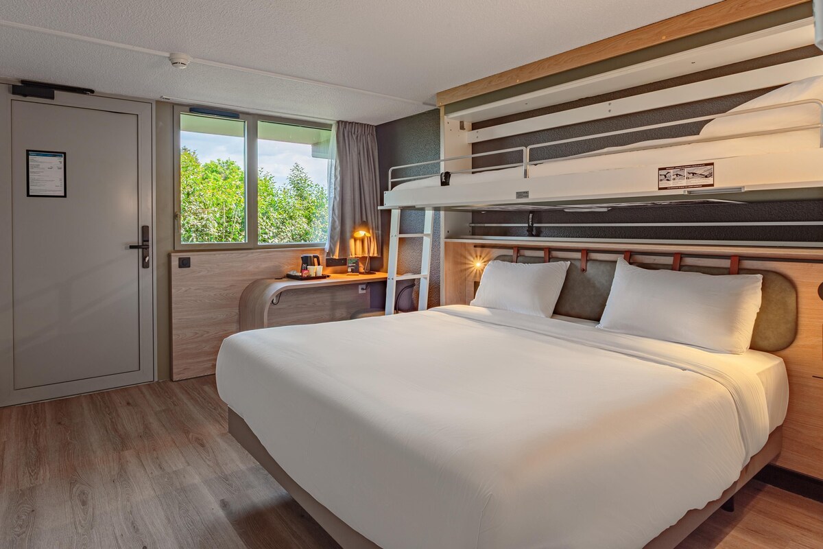 A hotel room with a bed, a desk with a lamp, a window with curtains, and a door.