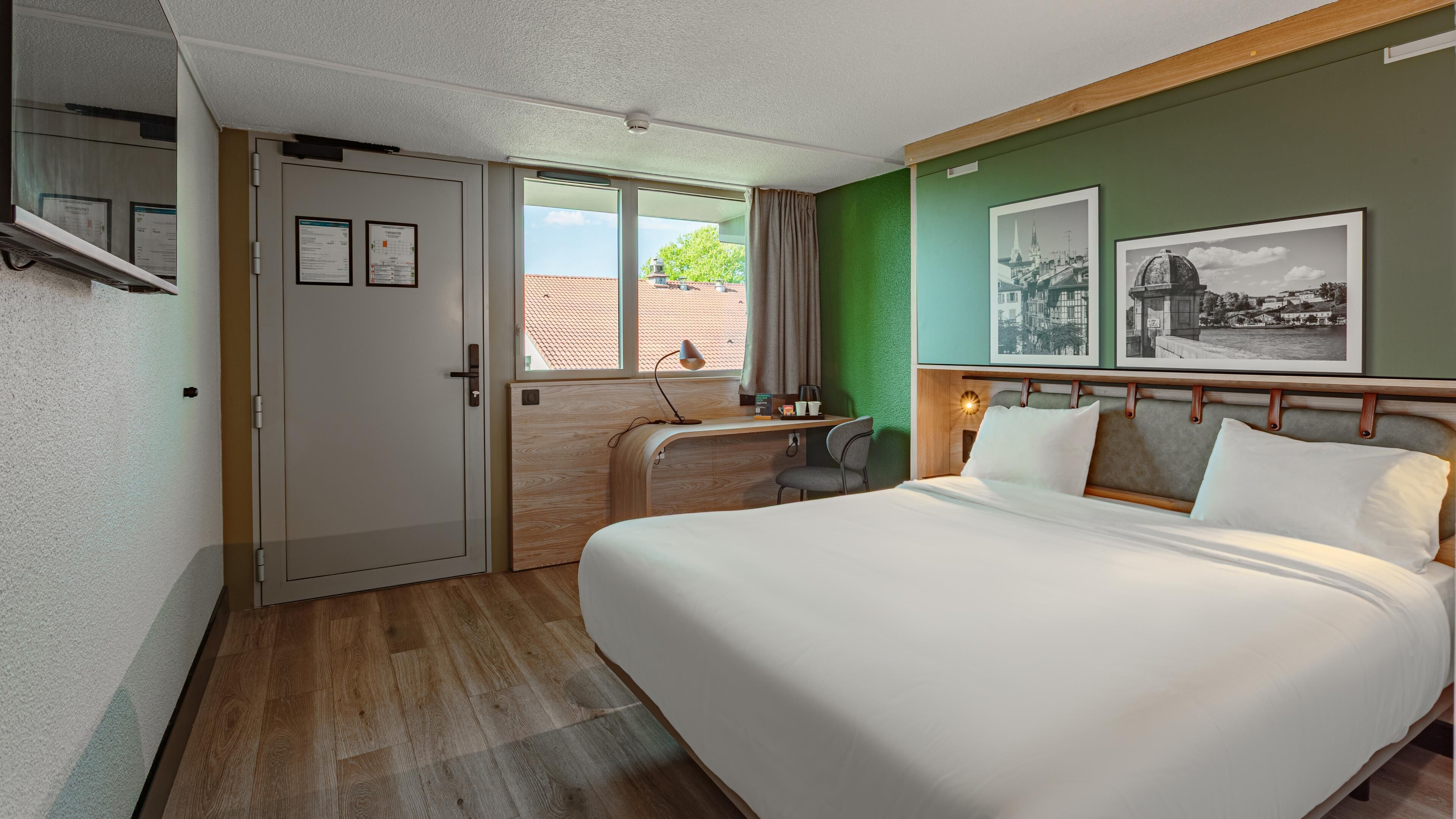 A hotel room with a bed, desk, chair, and a view of the city through a window.