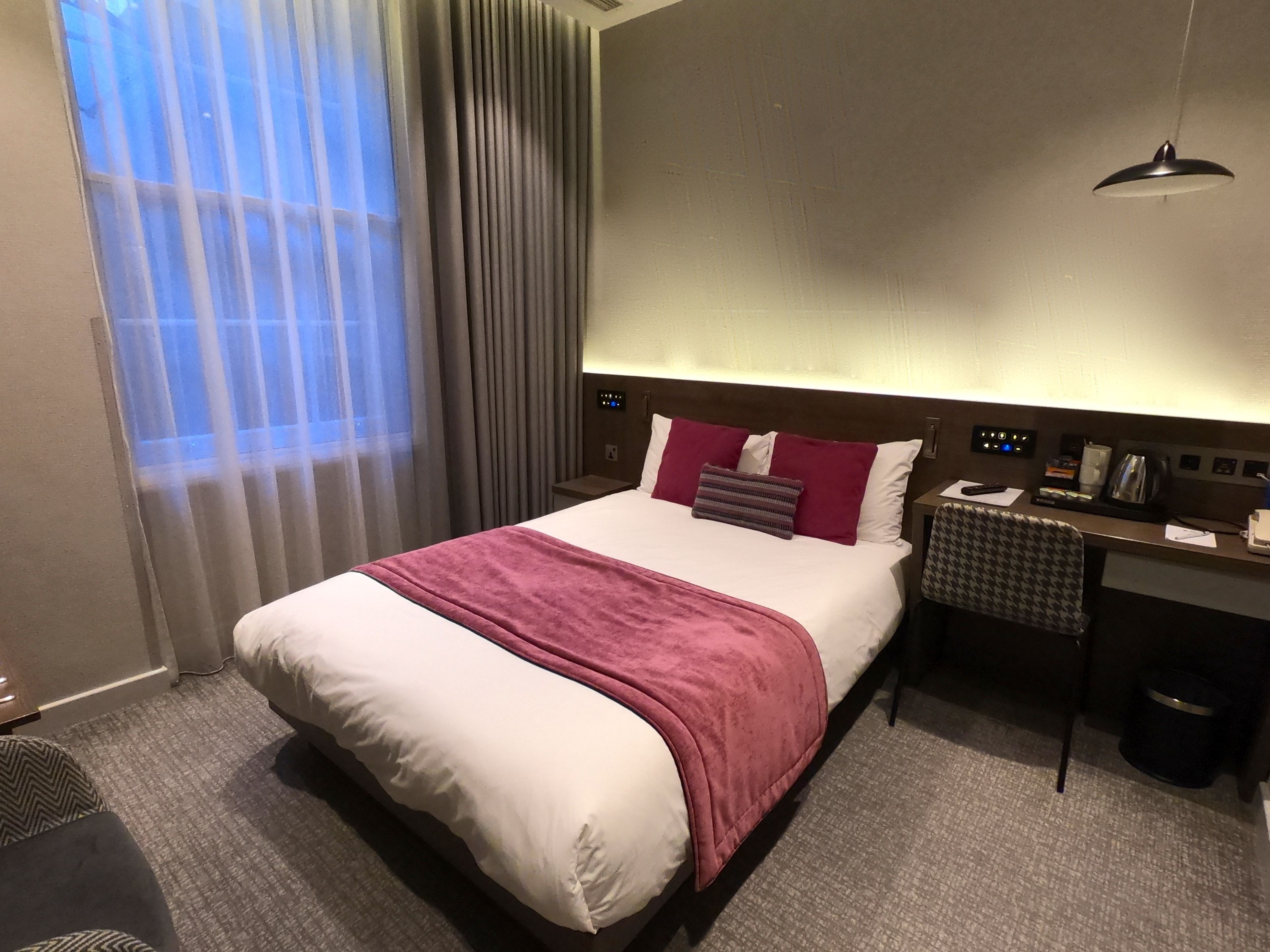Quarto de hotel com uma cama grande, uma escrivaninha com cadeira, uma lixeira e uma janela com cortinas.