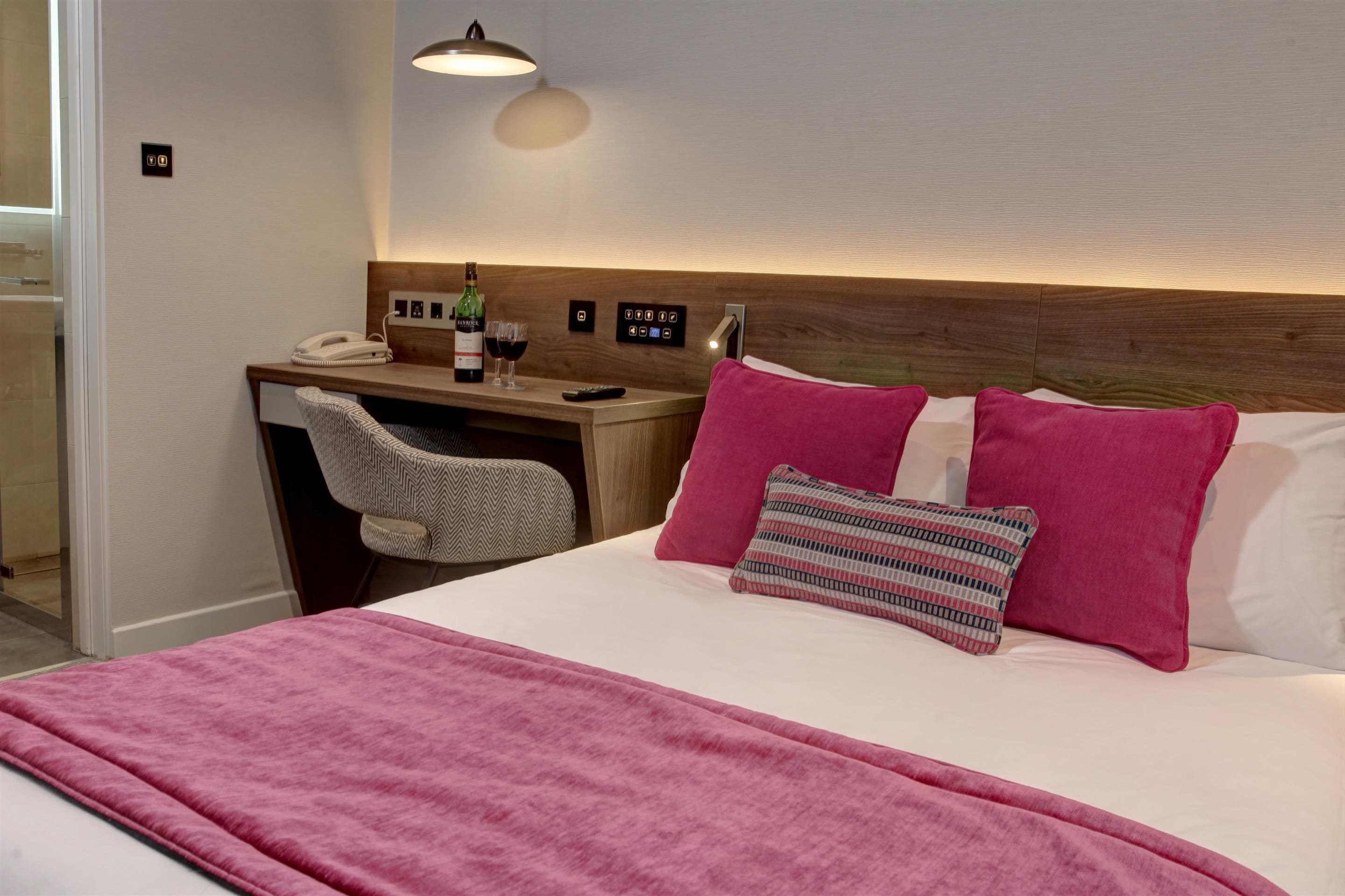 Habitación de hotel con cama, escritorio, teléfono, botella de vino y control remoto.