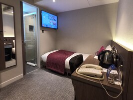 Et hotelværelse med seng, skrivebord med telefon, elkedel og et tv ophængt på væggen.