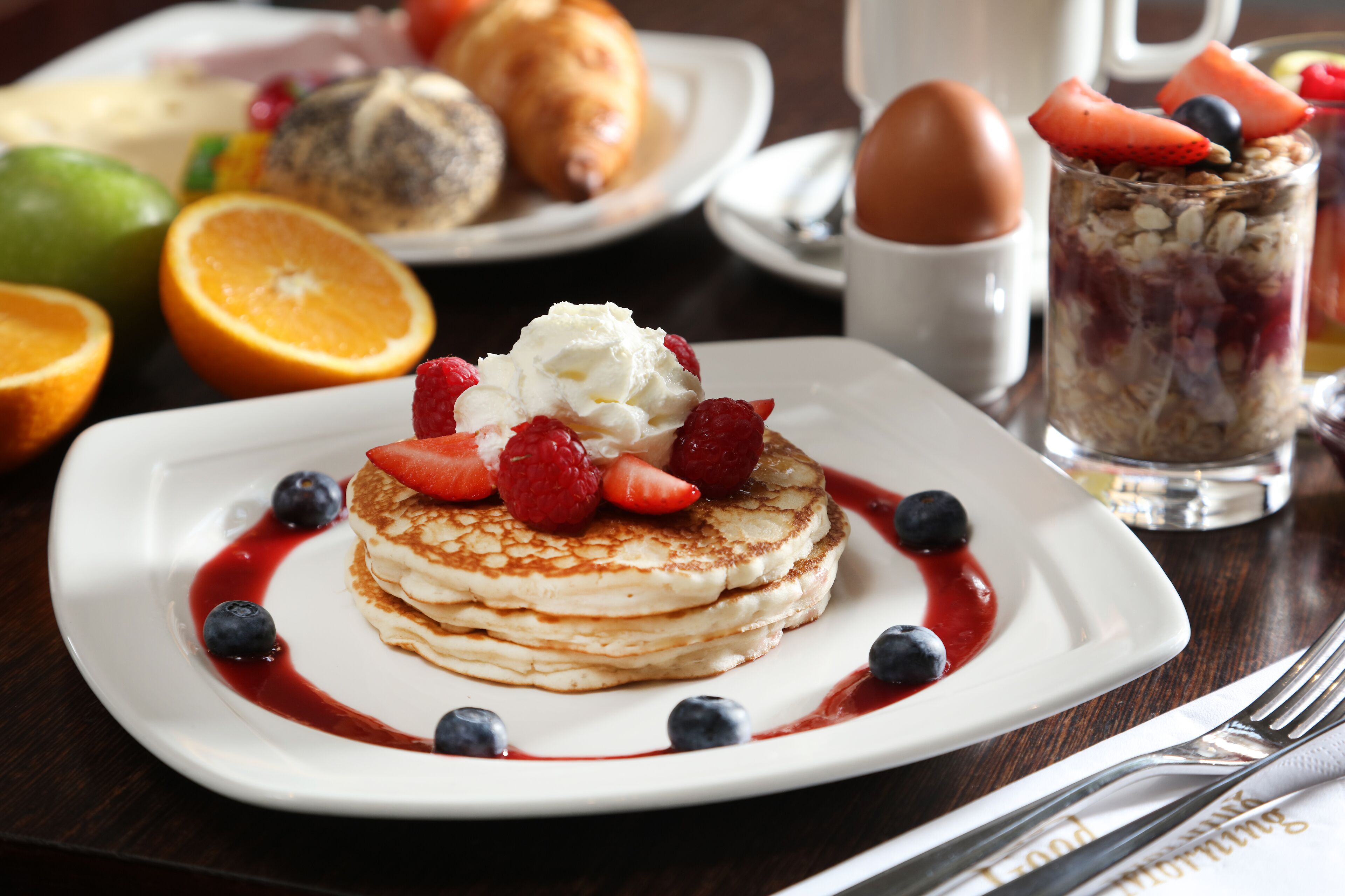 Un plato de desayuno con tortitas, fresas, arándanos, un croissant y un vaso de ensalada de frutas.