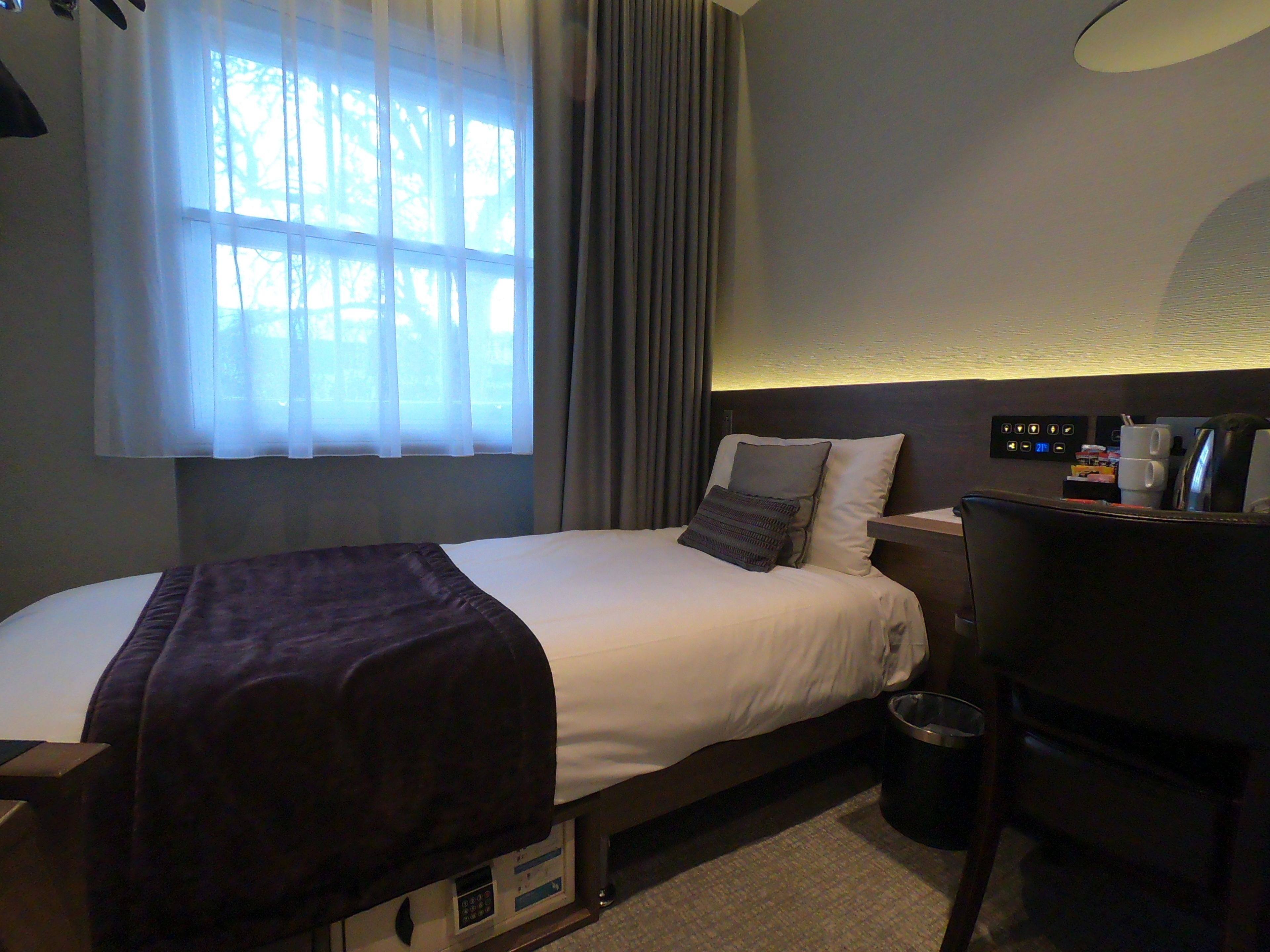 Habitación de hotel con cama, escritorio, silla, mesita de noche, lámpara, ventana con cortinas y panel de control en la pared.