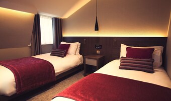 A hotel room with two beds, each with a red blanket and pillows, a bedside table with a lamp, and a window with curtains.