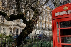 Eine rote Telefonzelle mit dem Schriftzug „TELEPHONE“, ein Baum mit Blüten und im Hintergrund ein Gebäude mit der Aufschrift „THE DELMERE HOTEL“.