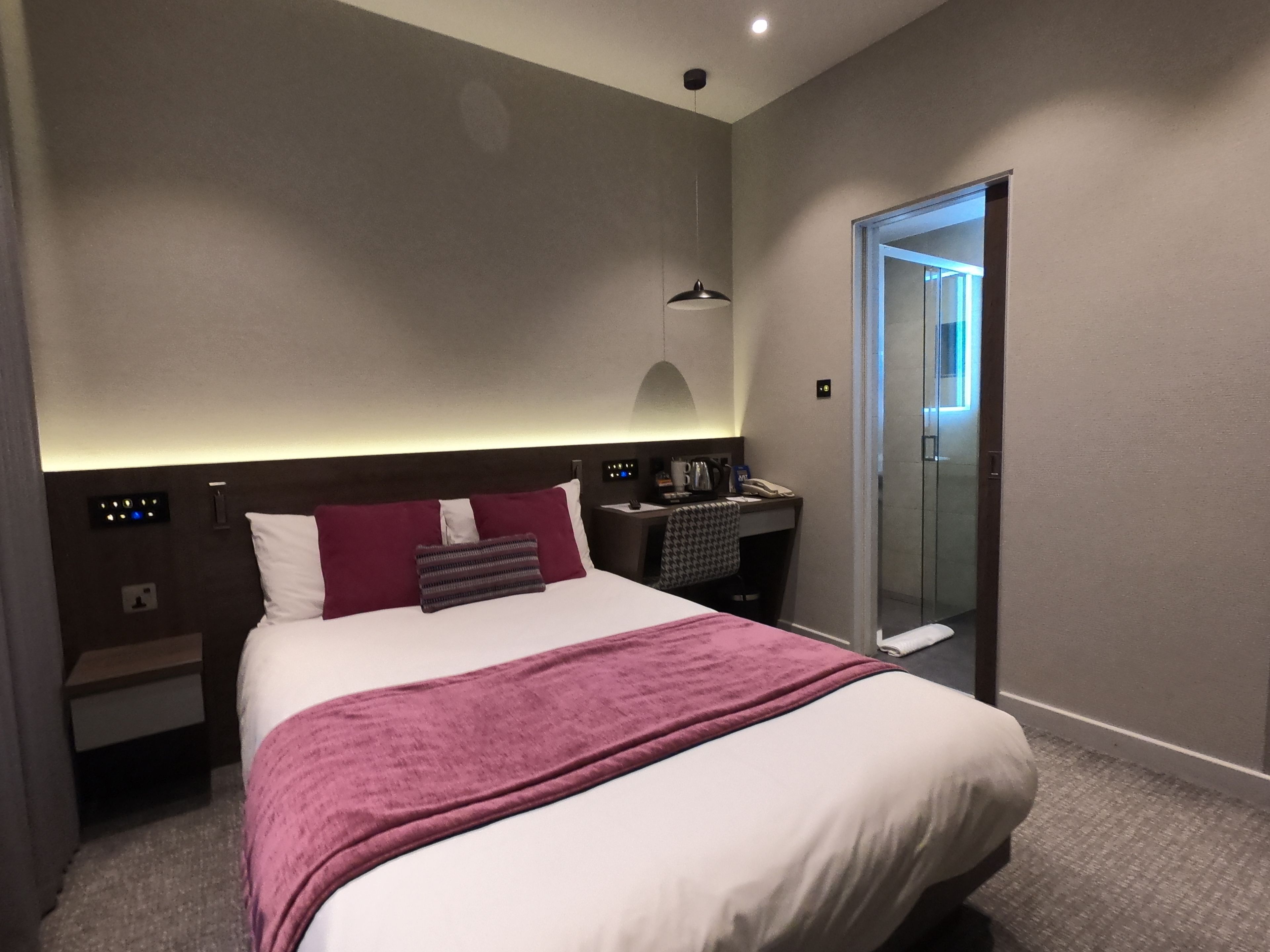 A modern hotel room with a large bed, a desk, a chair, a nightstand, a mirror, and a door leading to a bathroom.