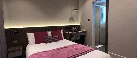 A modern hotel room with a large bed, a desk, a chair, a nightstand, a mirror, and a door leading to a bathroom.
