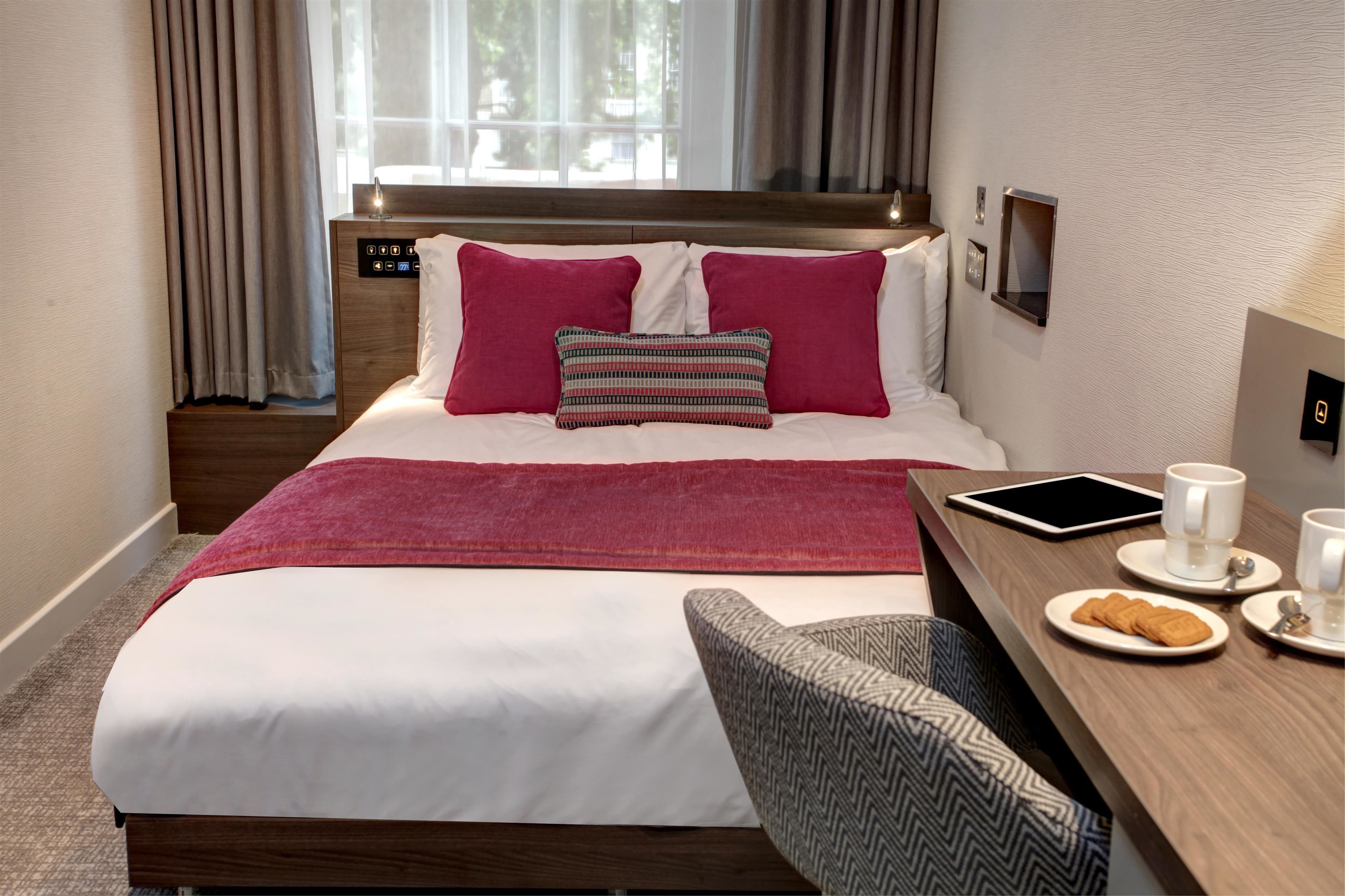 A hotel room with a bed, a bedside table with a tablet, a chair, and a breakfast setup with pastries and coffee cups.
