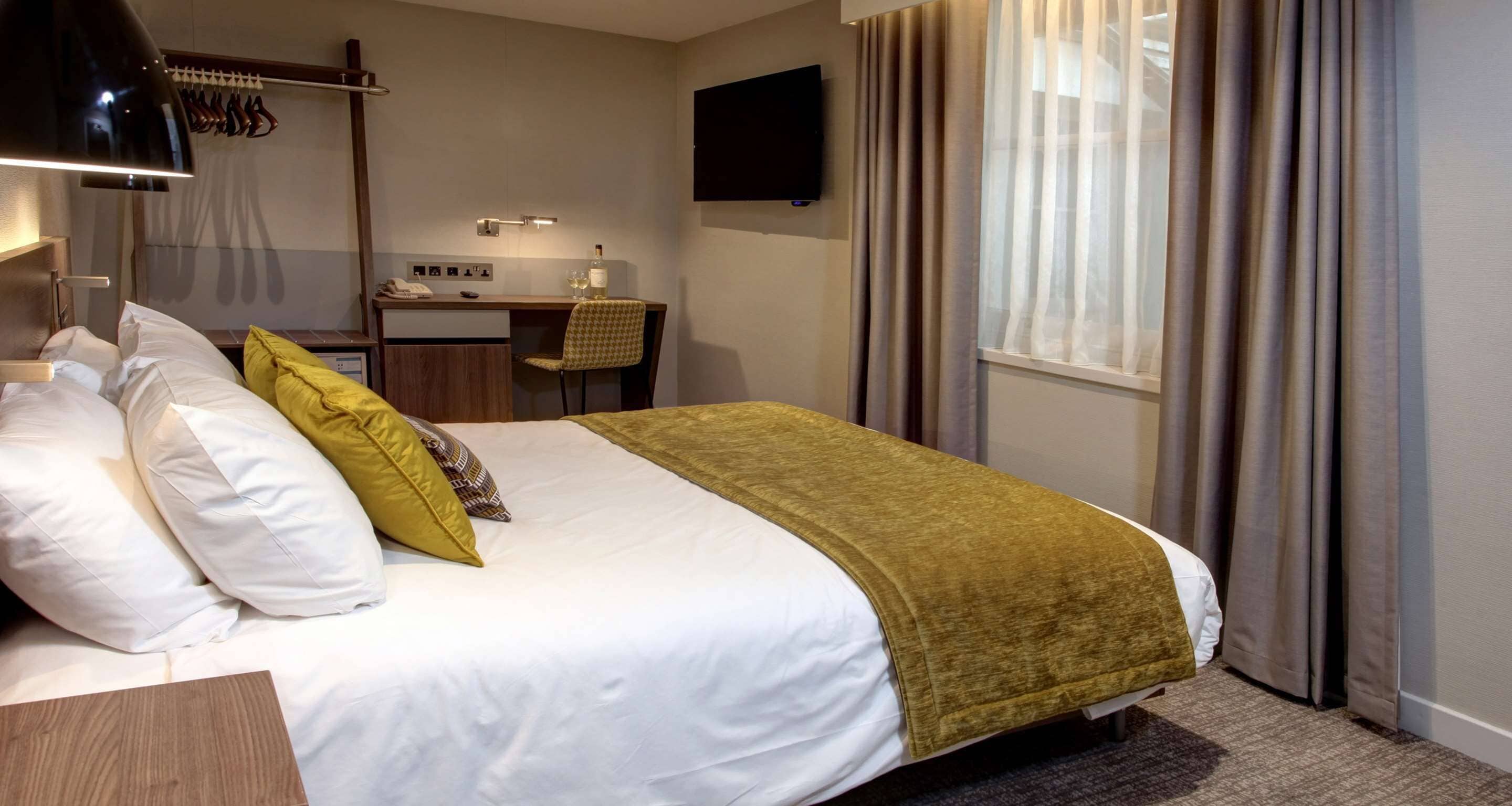 Habitación de hotel con cama, escritorio, silla, televisor y ventana con cortinas.