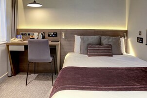 Ein Hotelzimmer mit Bett, Schreibtisch, Stuhl, Telefon, Kaffeemaschine und Bedienfeld an der Wand.