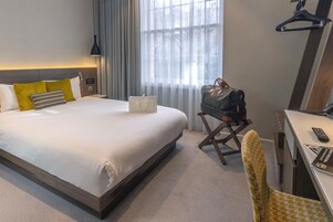 A hotel room with a bed, a desk, a chair, a hanging coat rack, and a window with curtains.