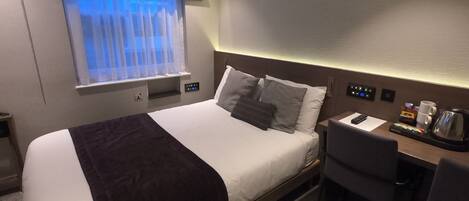 A hotel room with a bed, a desk, a chair, a window with curtains, a bedside table with a coffee maker, and a control panel.