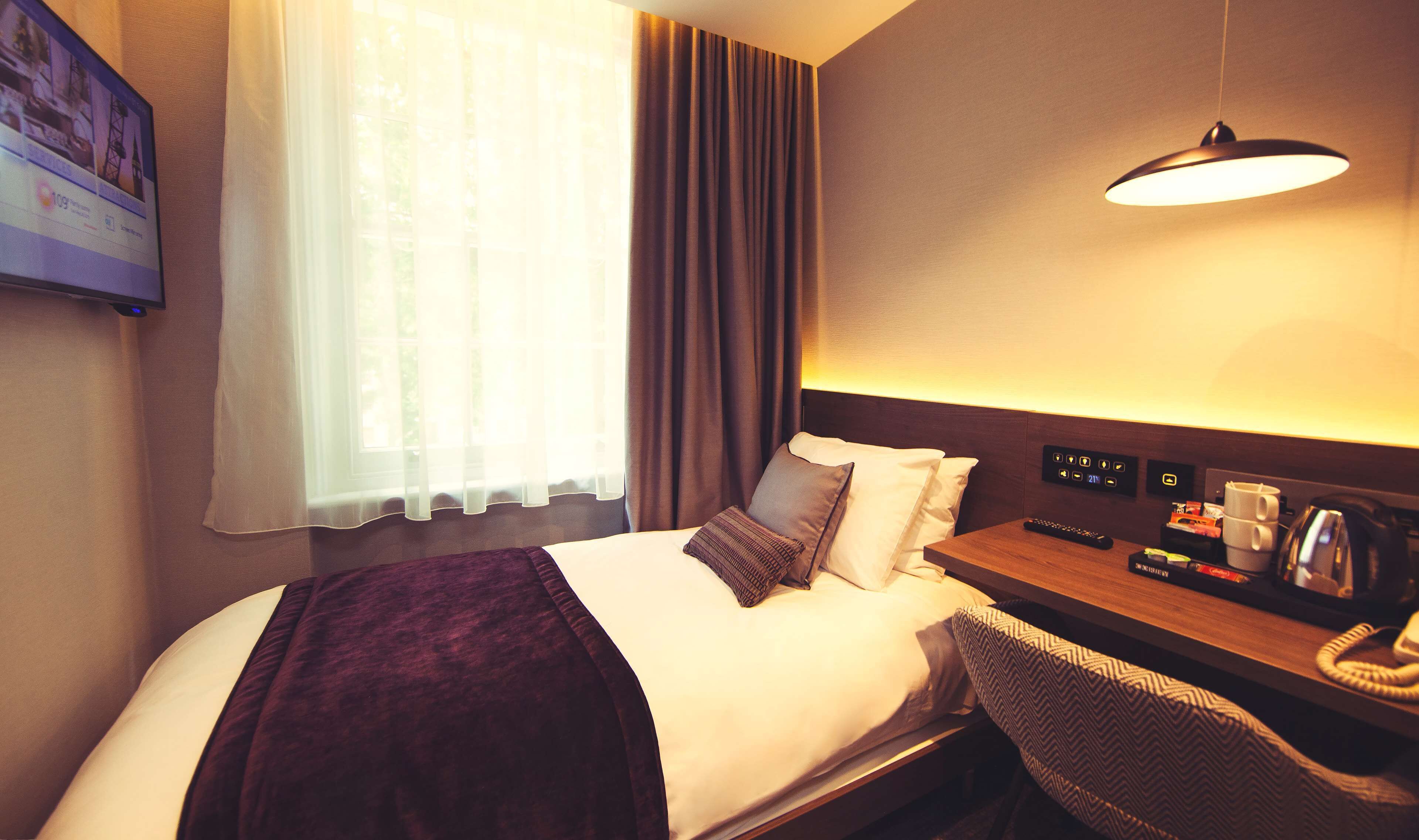 Ein Hotelzimmer mit einem Bett, einem Schreibtisch mit Lampe, einem Fernseher und einem Fenster mit Vorhängen.