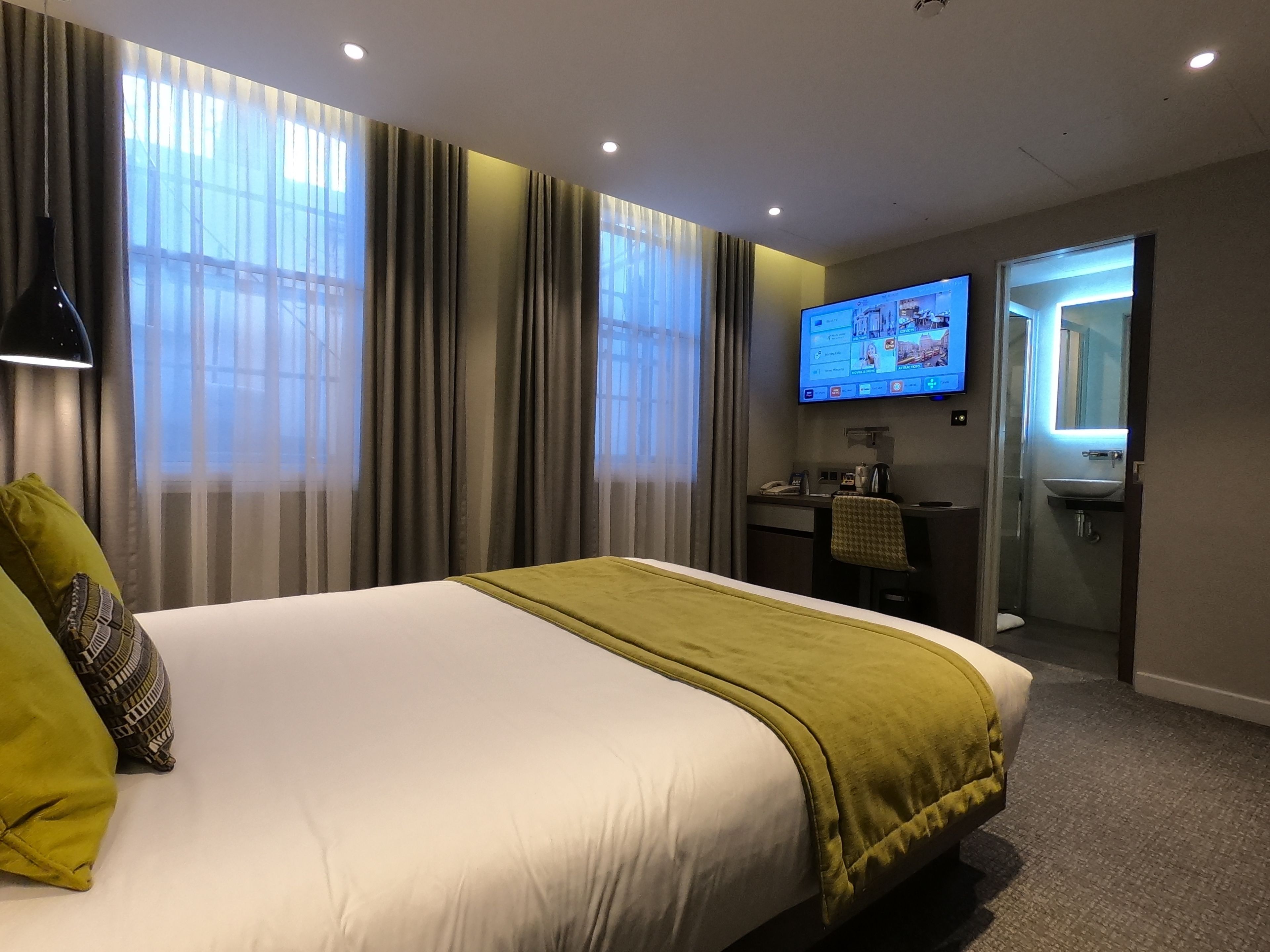 Habitación de hotel con una cama grande, un escritorio con una computadora, un televisor y un baño a la vista a través de una puerta abierta.